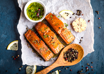 Cooked salmon fillets on a paper, accompanied by lemon slices, peppercorns, a wooden spoon, and a small bowl of green sauce.