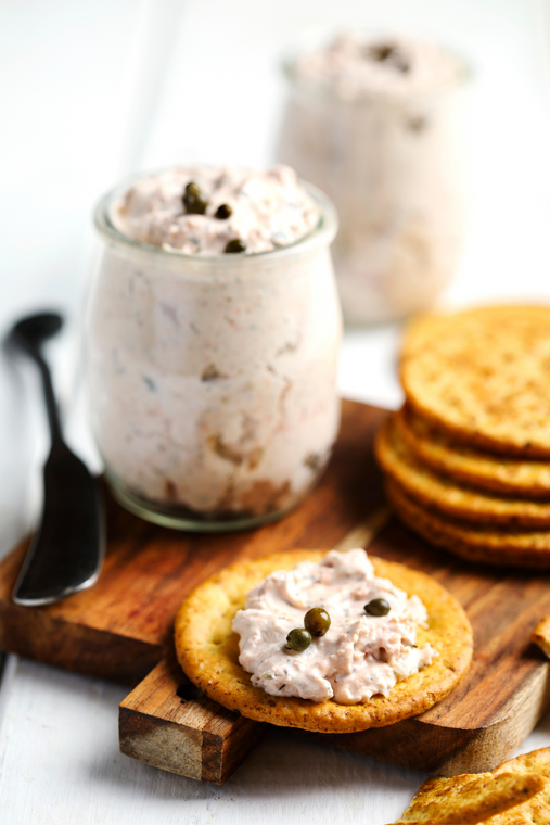A jar of smoked fish dip on a wooden board next to crackers and a knife, with a blurred background.