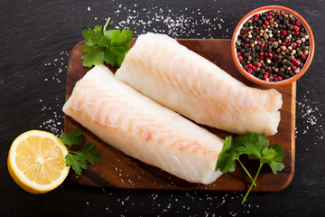 Two fillets of golden tilefish on a wooden cutting board with lemon slices and herbs on the side, and a small bowl of seasonings in the background. 